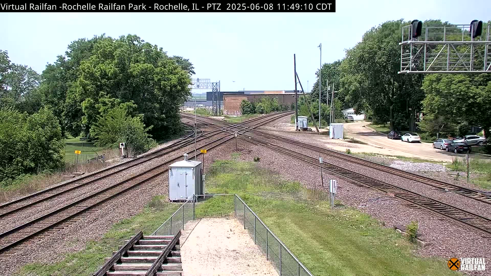 A sunny day reveals a railway crossing with several train tracks intersecting, surrounded by green trees and a few parked cars in the distance.