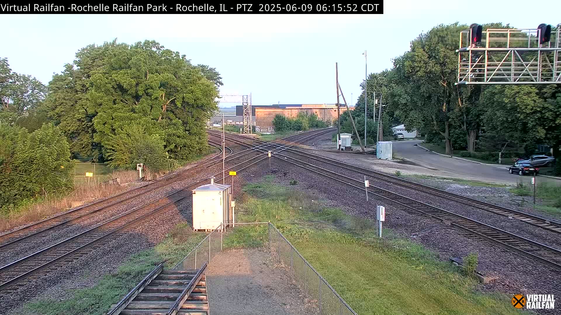 A railroad crossing with multiple tracks is shown under a partly cloudy sky, surrounded by trees and grass.