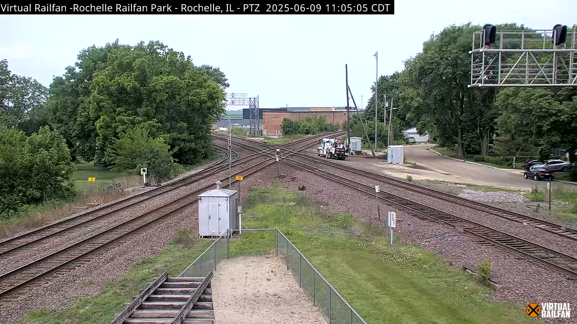 A railroad crossing with multiple tracks is seen on a partly sunny day, with a service truck parked near the intersection and vehicles parked along a nearby road.
