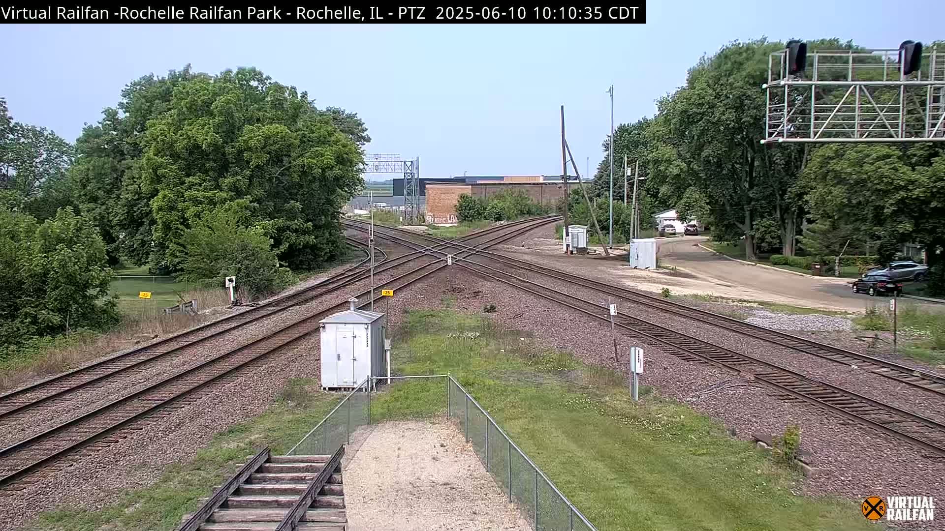 A sunny outdoor view of a railway crossing with several train tracks intersecting, surrounded by green trees and grass, and a small building near the tracks.