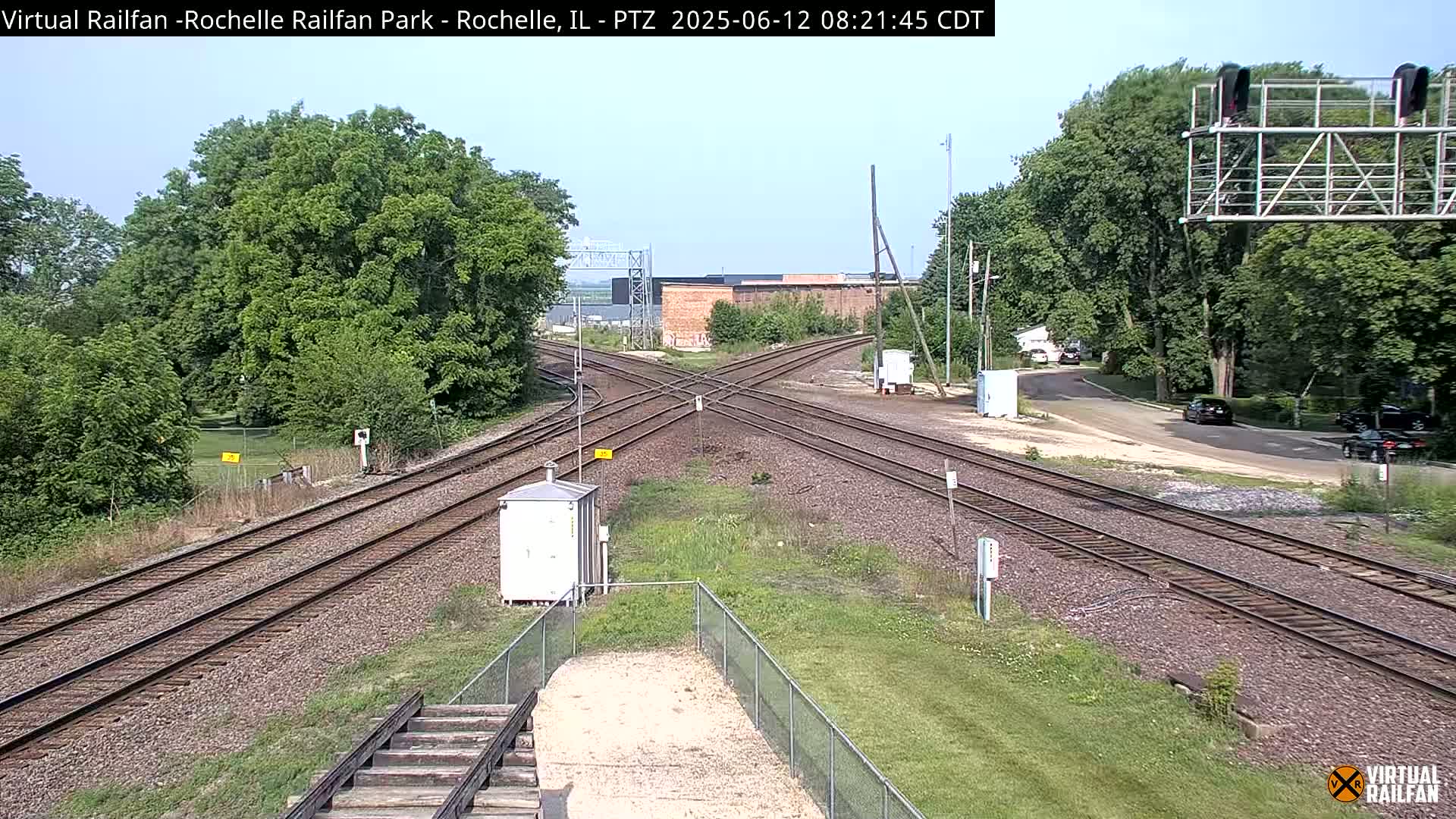A sunny day reveals a railroad crossing with multiple tracks intersecting, surrounded by green trees and grass.
