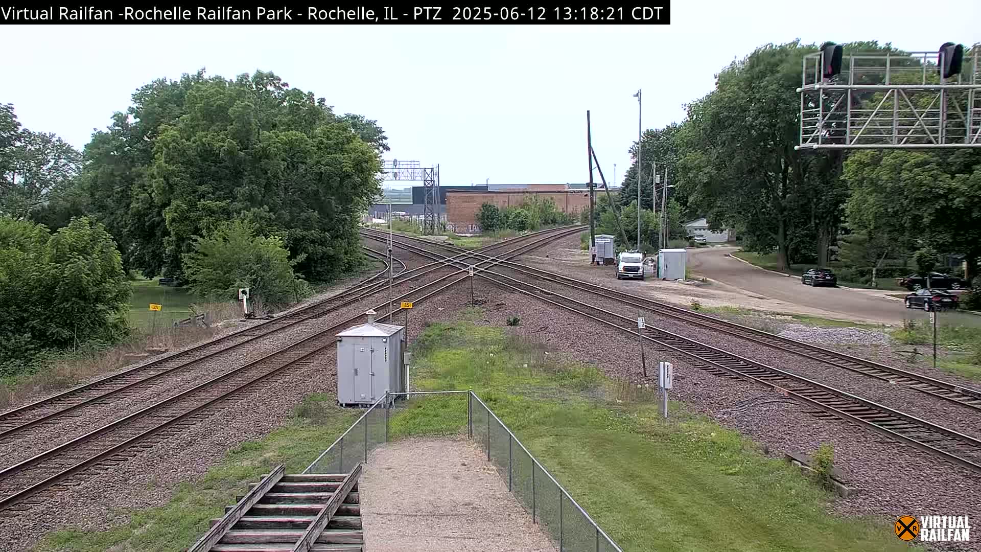 A high-angle, long shot of a railroad crossing with several train tracks intersecting, under a mostly cloudy sky, surrounded by green trees and grass.
