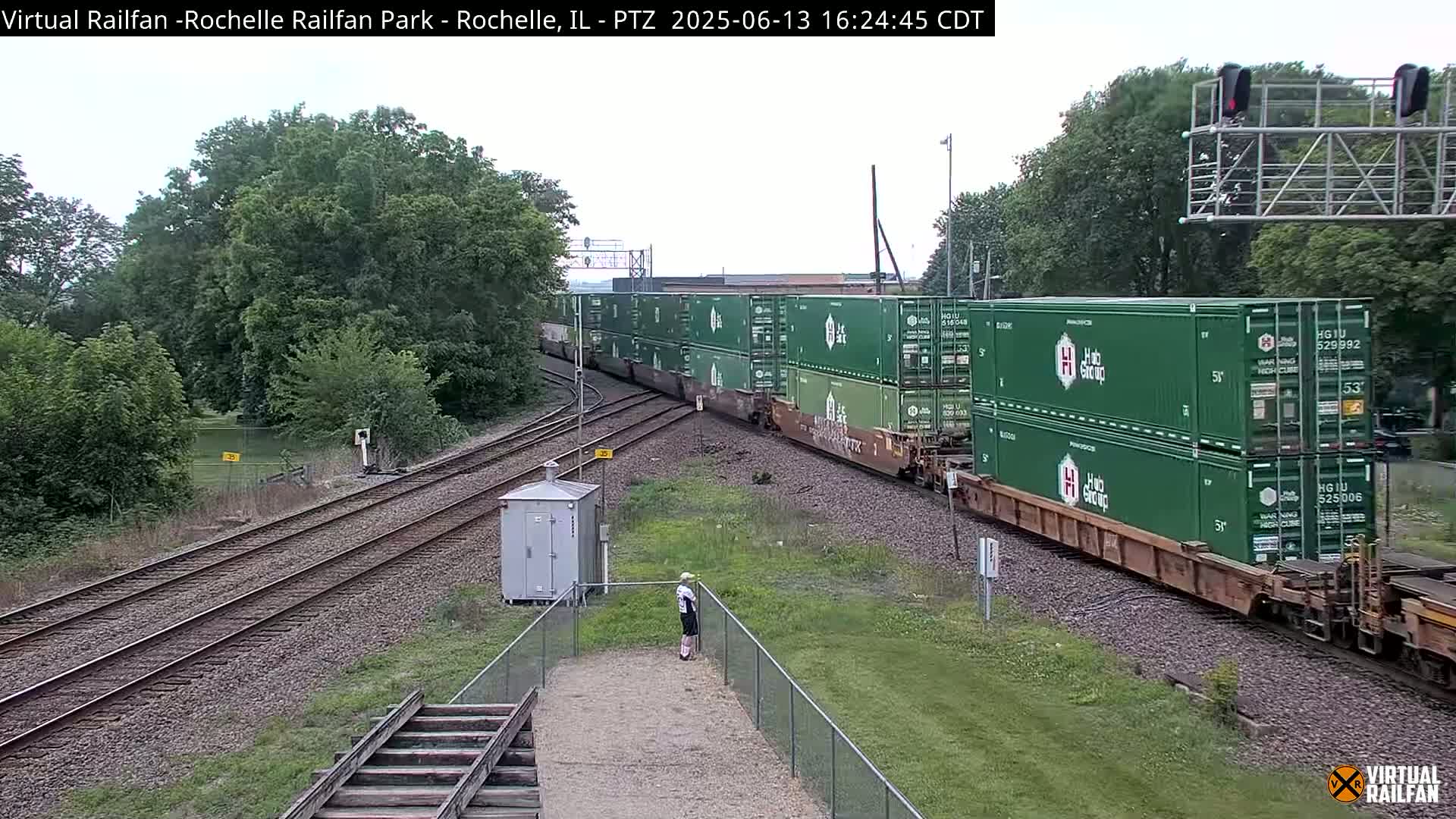 A long freight train carrying many green shipping containers travels along railroad tracks on an overcast day, past a person standing near a chain-link fence.