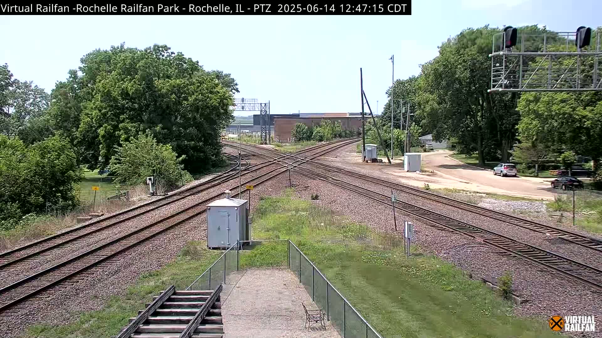 A high-angle view of a railroad crossing with intersecting tracks, surrounded by green trees and grass under a partly sunny sky.