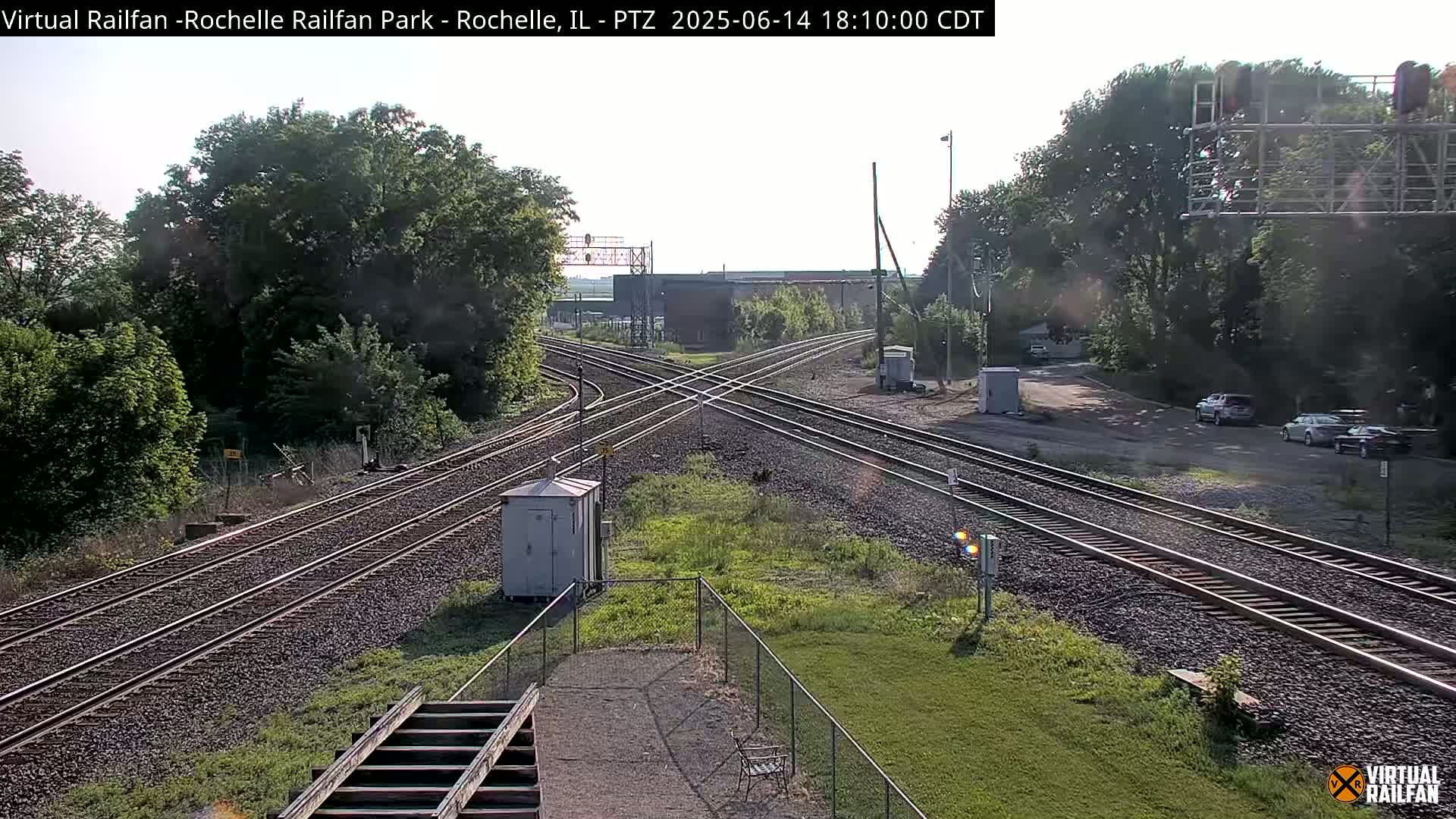 A high-angle view of a railroad crossing with intersecting tracks, surrounded by green trees and grass under a partly sunny sky.