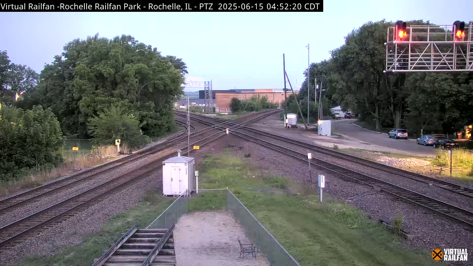 A railroad crossing with several train tracks intersecting under a twilight sky, surrounded by lush green trees and vegetation.