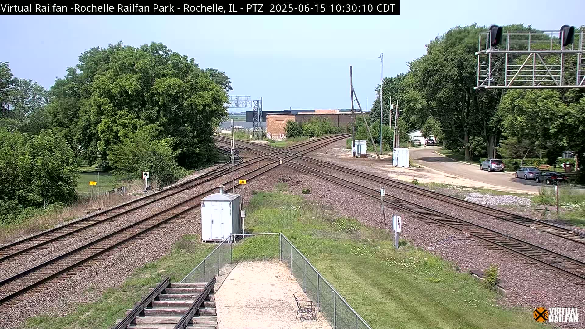 A high-angle view of a sunny railroad crossing with several tracks intersecting, surrounded by green trees and grass, with a few cars visible on a nearby road.
