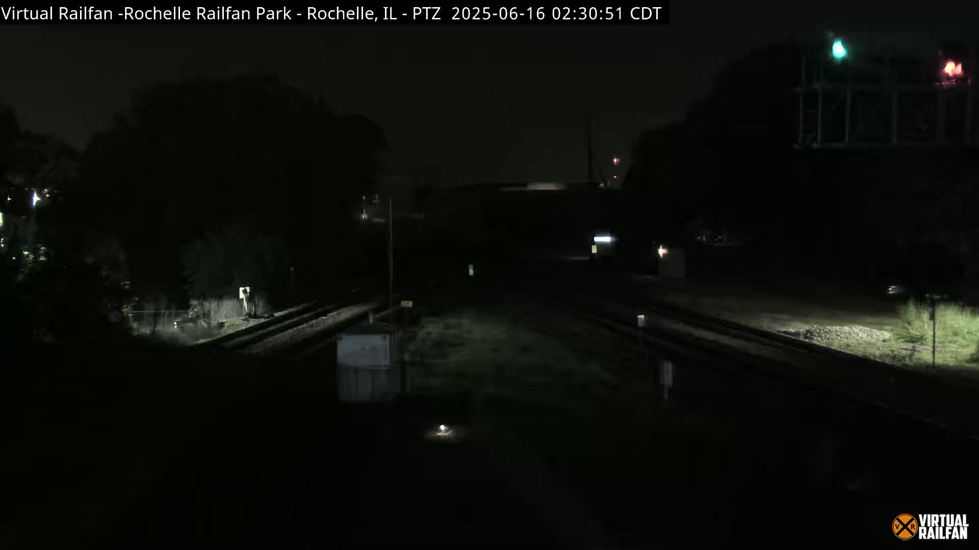 A nighttime, dark view of railroad tracks intersecting, with minimal ambient light and a structure with illuminated signals in the upper right corner.