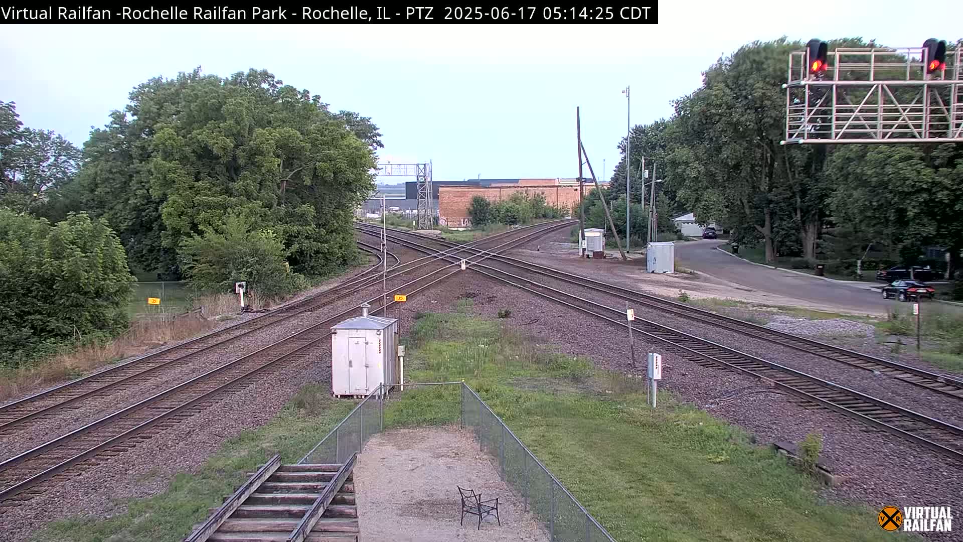 A railroad crossing with several sets of tracks, surrounded by green vegetation under a partly cloudy sky.