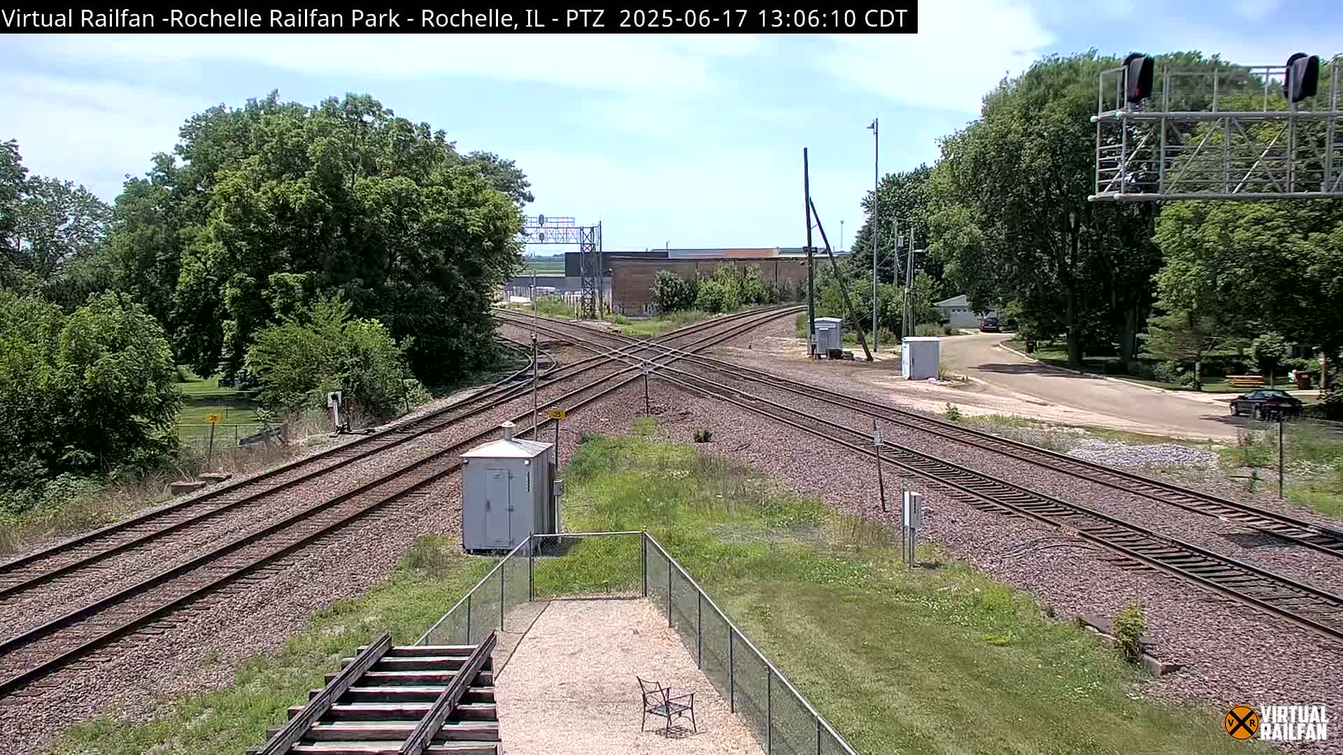 A sunny day shows a railroad crossing with multiple train tracks intersecting, surrounded by green trees and grassy areas.