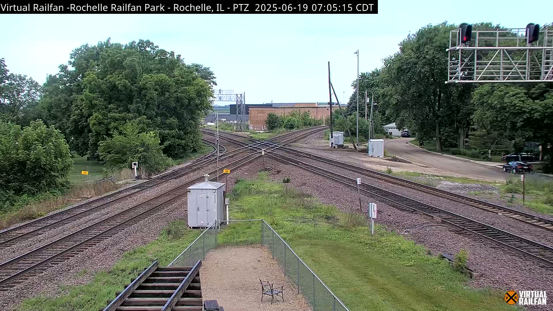 A mostly sunny view of a railroad crossing with multiple tracks, surrounded by green trees and grass.