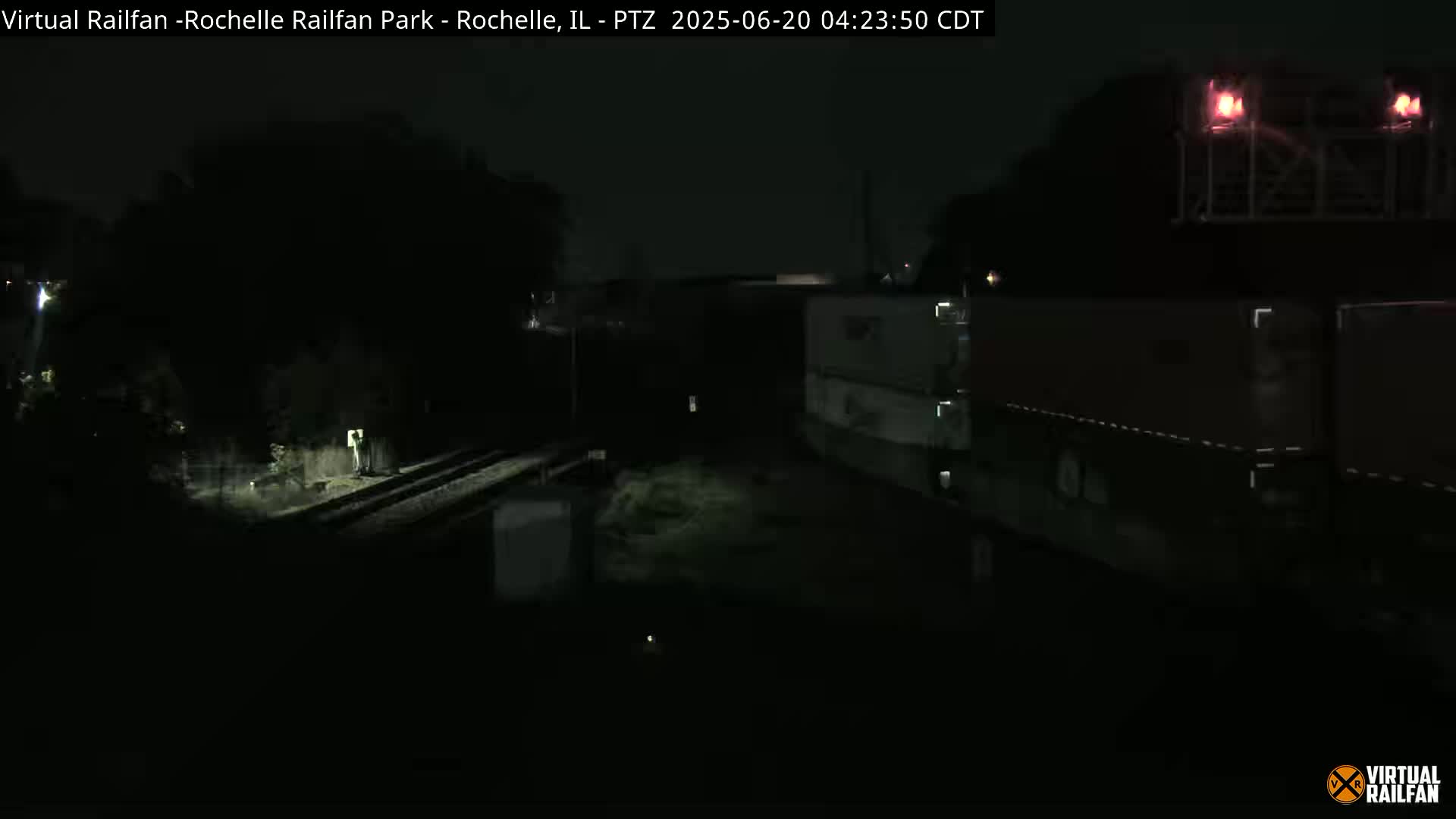 A nighttime, dimly lit view of railroad tracks and a train consisting of several dark cargo containers.