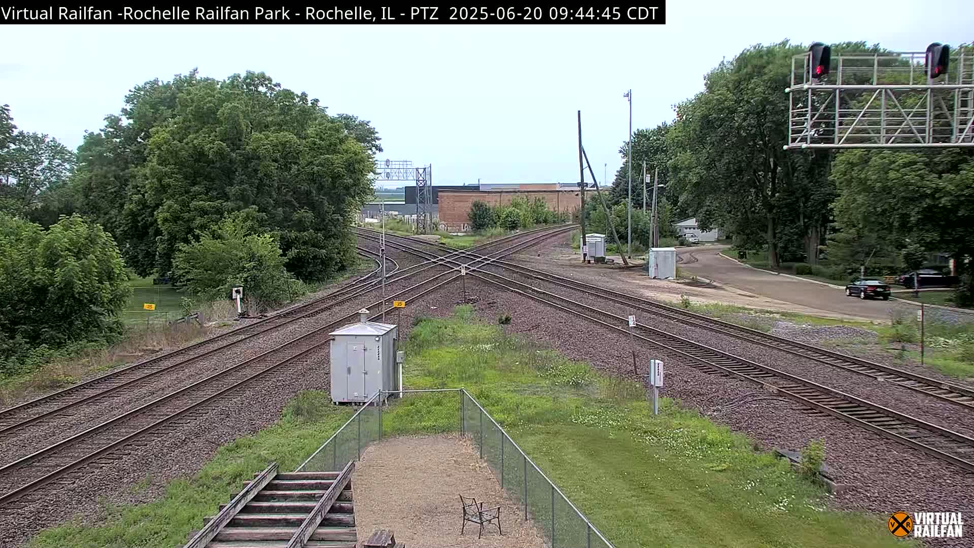 A railroad crossing with multiple tracks under a partly cloudy sky, surrounded by green trees and grass.