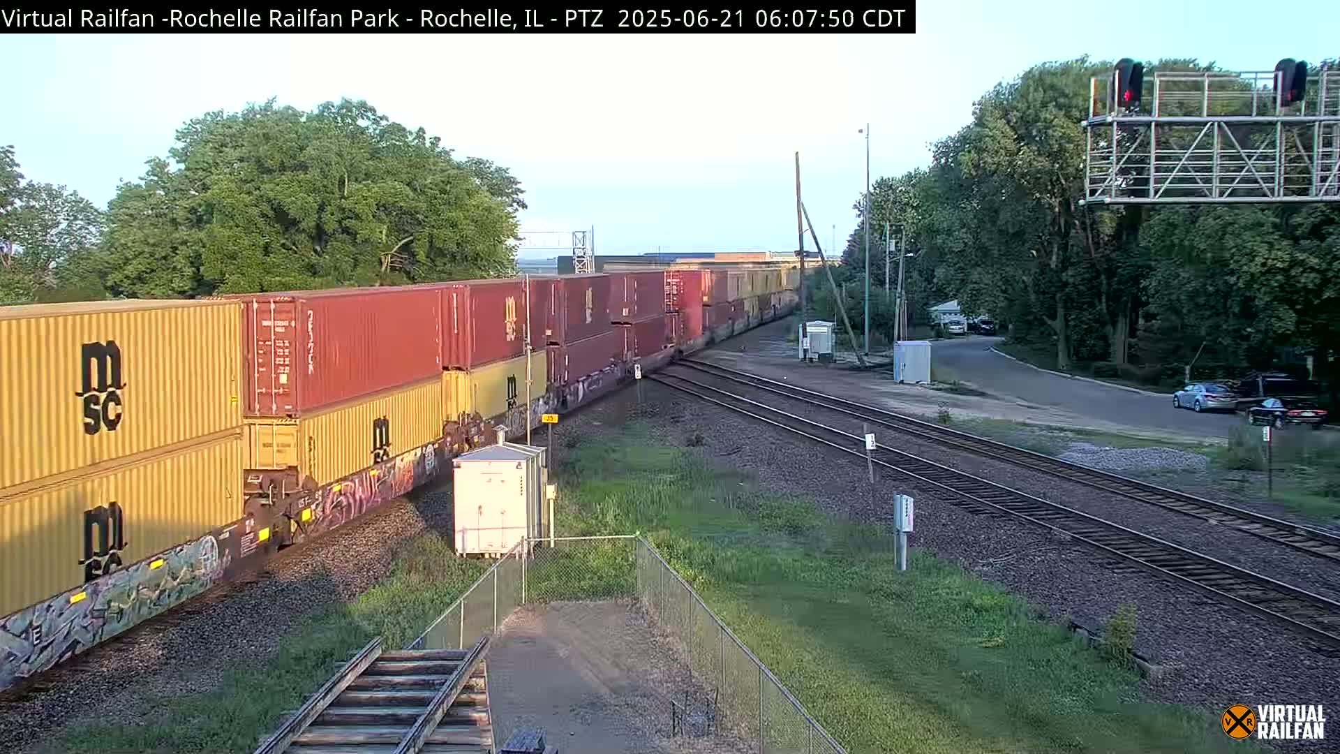A long freight train carrying multiple shipping containers moves past a roadway on a clear day.