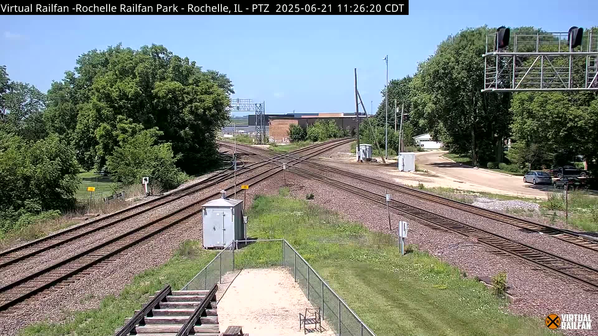 A sunny day reveals a railroad crossing with intersecting tracks, surrounded by lush green trees and grassy areas.