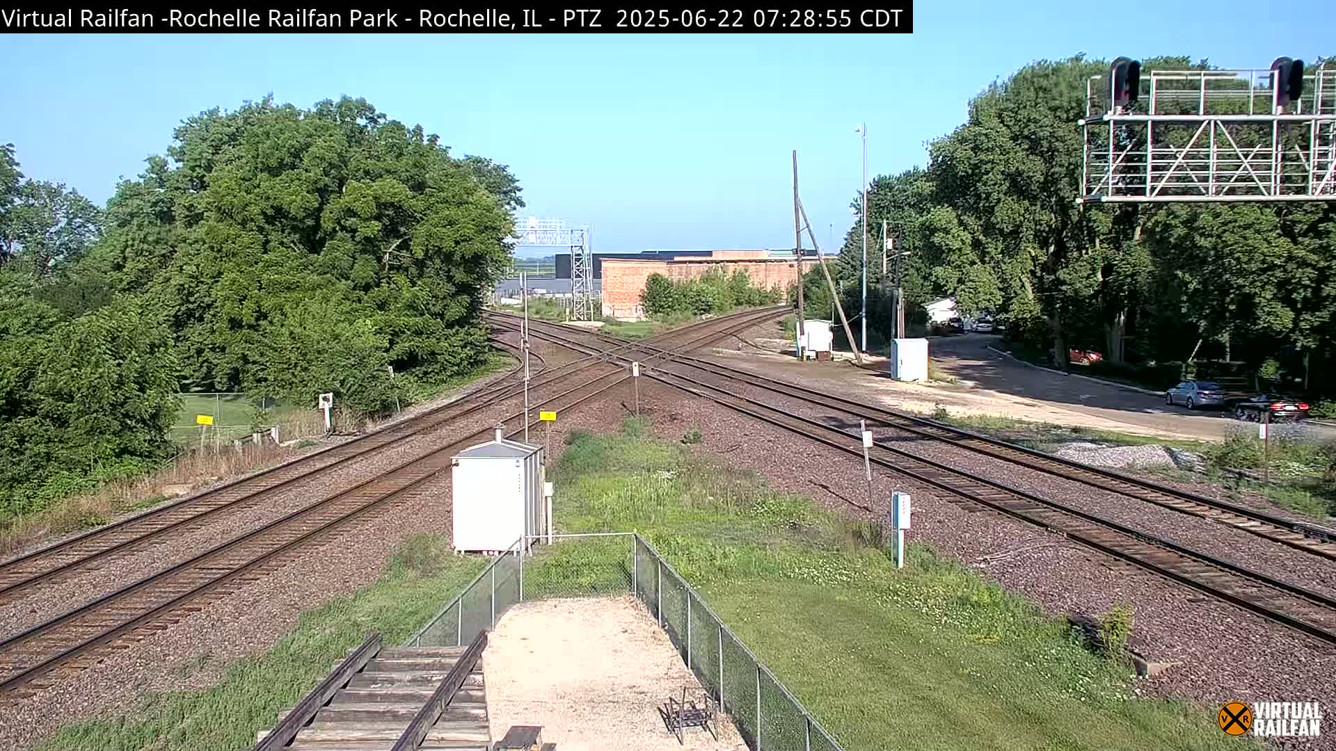 A sunny day shows a railway crossing with multiple tracks, surrounded by green trees and grass, and a few cars visible in the distance.