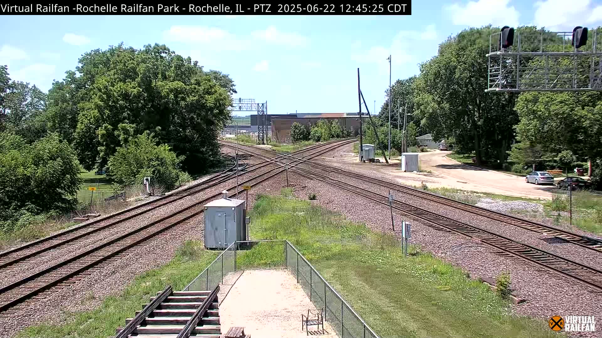 A sunny day shows a railroad crossing with multiple tracks, surrounded by green trees and vegetation.