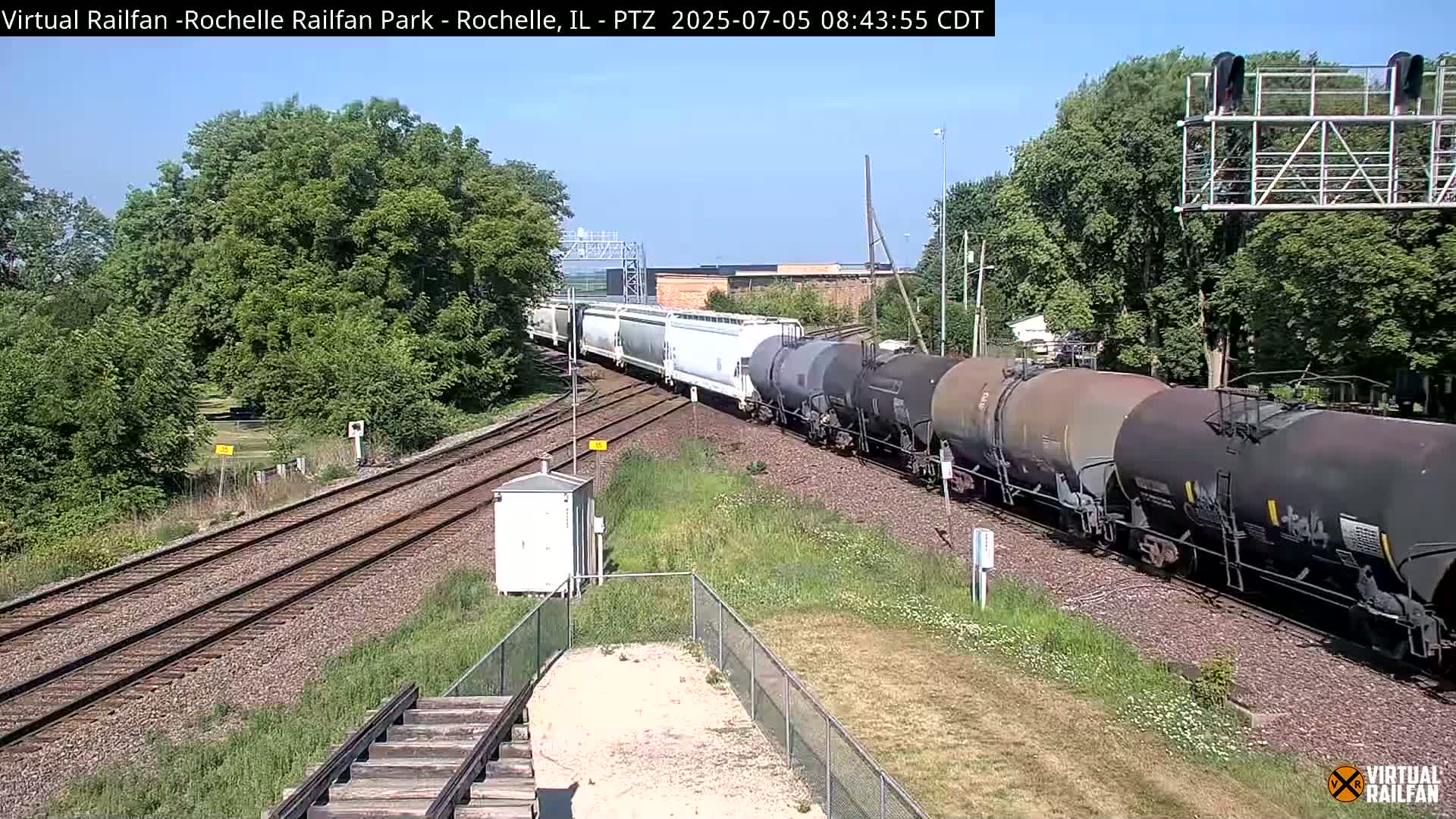 A long freight train, consisting of both covered hopper and tank cars, moves along railroad tracks on a sunny day, surrounded by lush green trees.