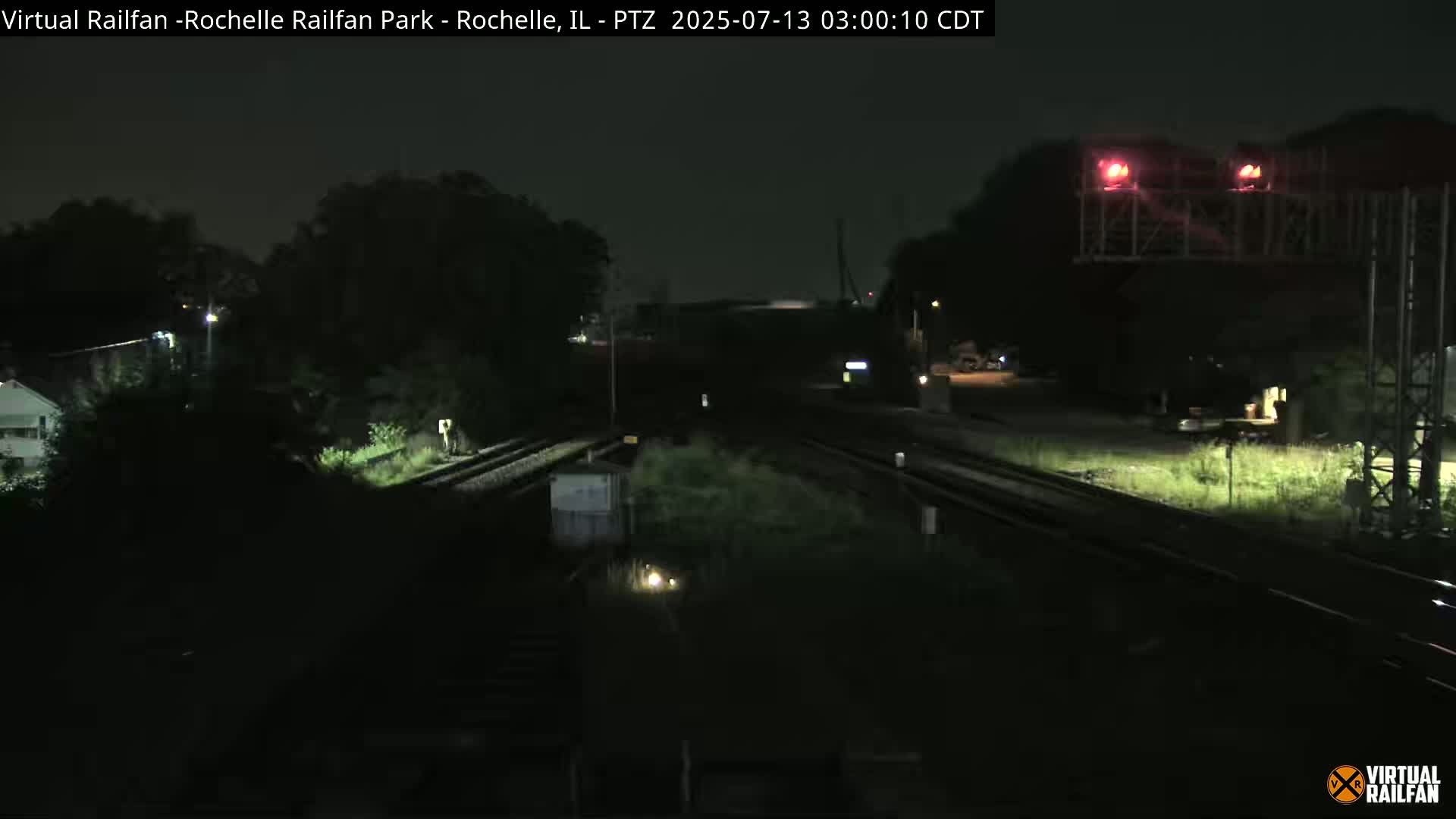 A nighttime view of a dark, mostly empty train yard with railroad tracks and some vegetation.
