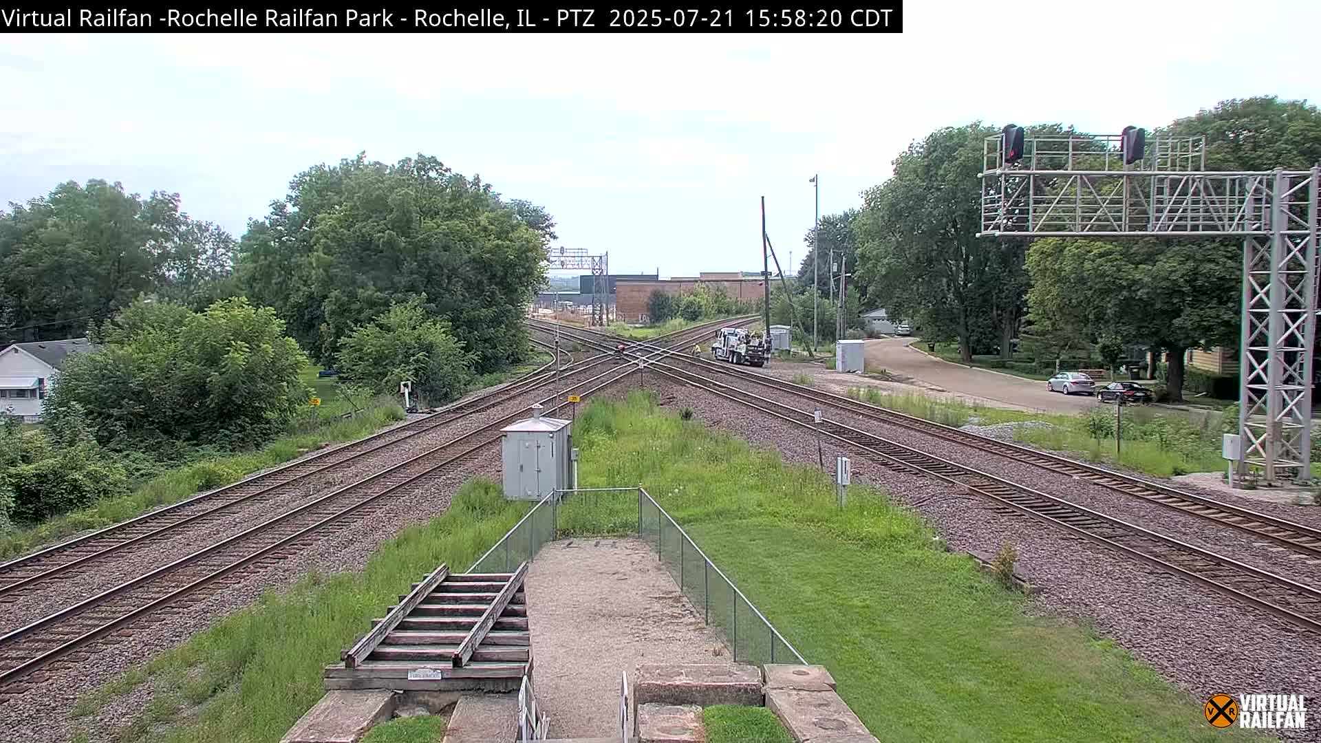 A mostly sunny view of a railroad crossing with several train tracks, a small service vehicle, and surrounding green vegetation.