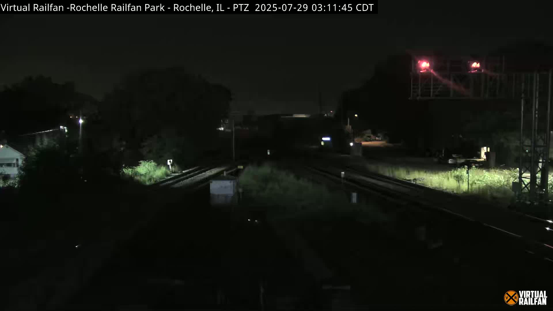 A nighttime view of railroad tracks intersecting, with some vegetation and a building visible in the distance under a dark sky.