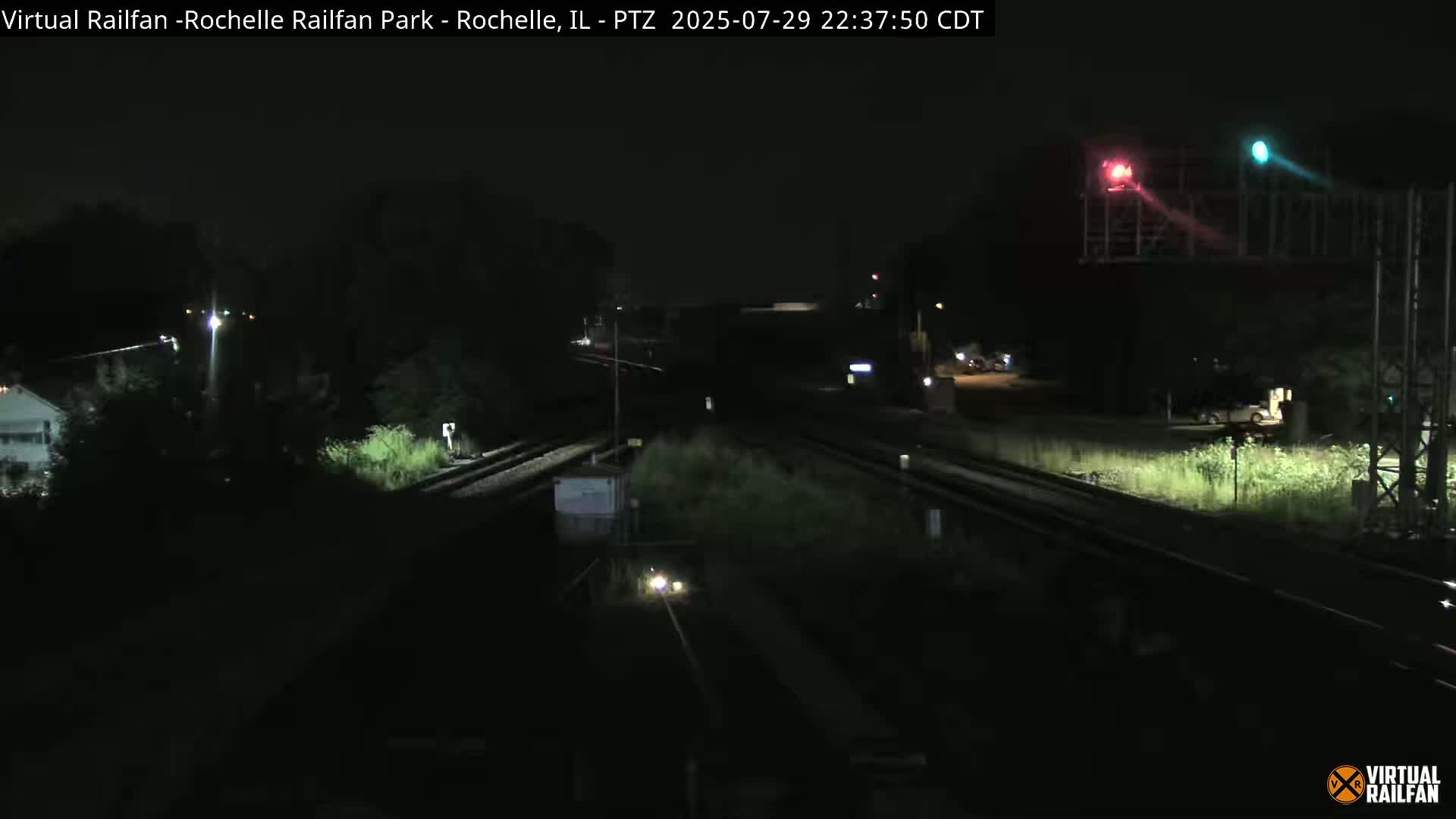A nighttime view of railroad tracks intersecting, with some vegetation and a building visible in the distance under a dark sky.