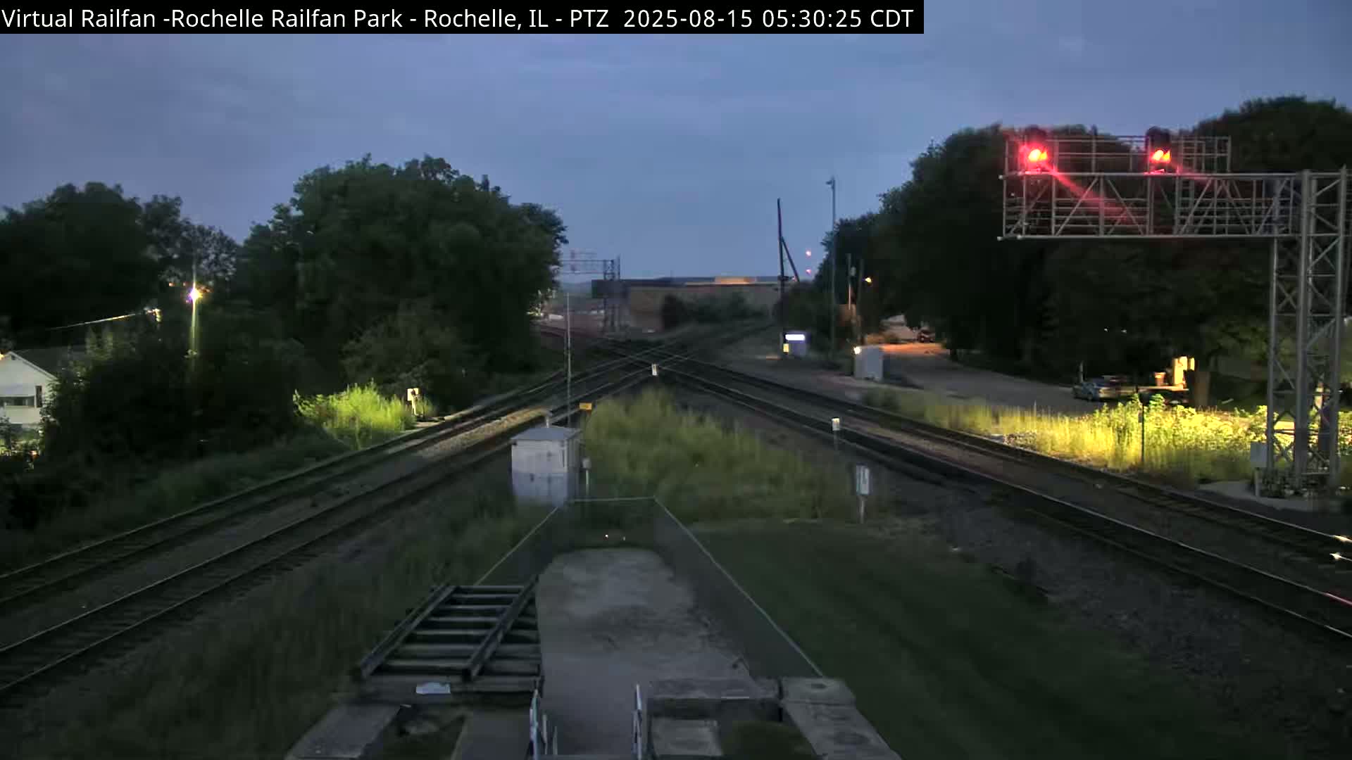 A nighttime view of railroad tracks intersecting, with some vegetation and a building visible in the distance under a dark sky.