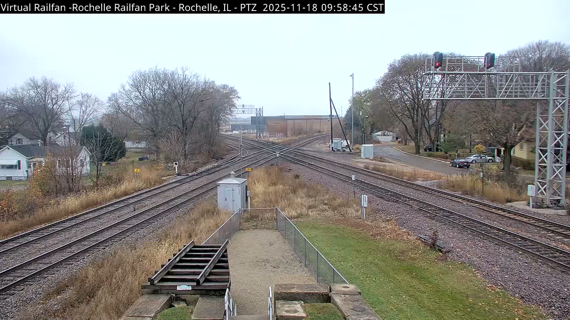 A nighttime view of railroad tracks intersecting, with some vegetation and a building visible in the distance under a dark sky.