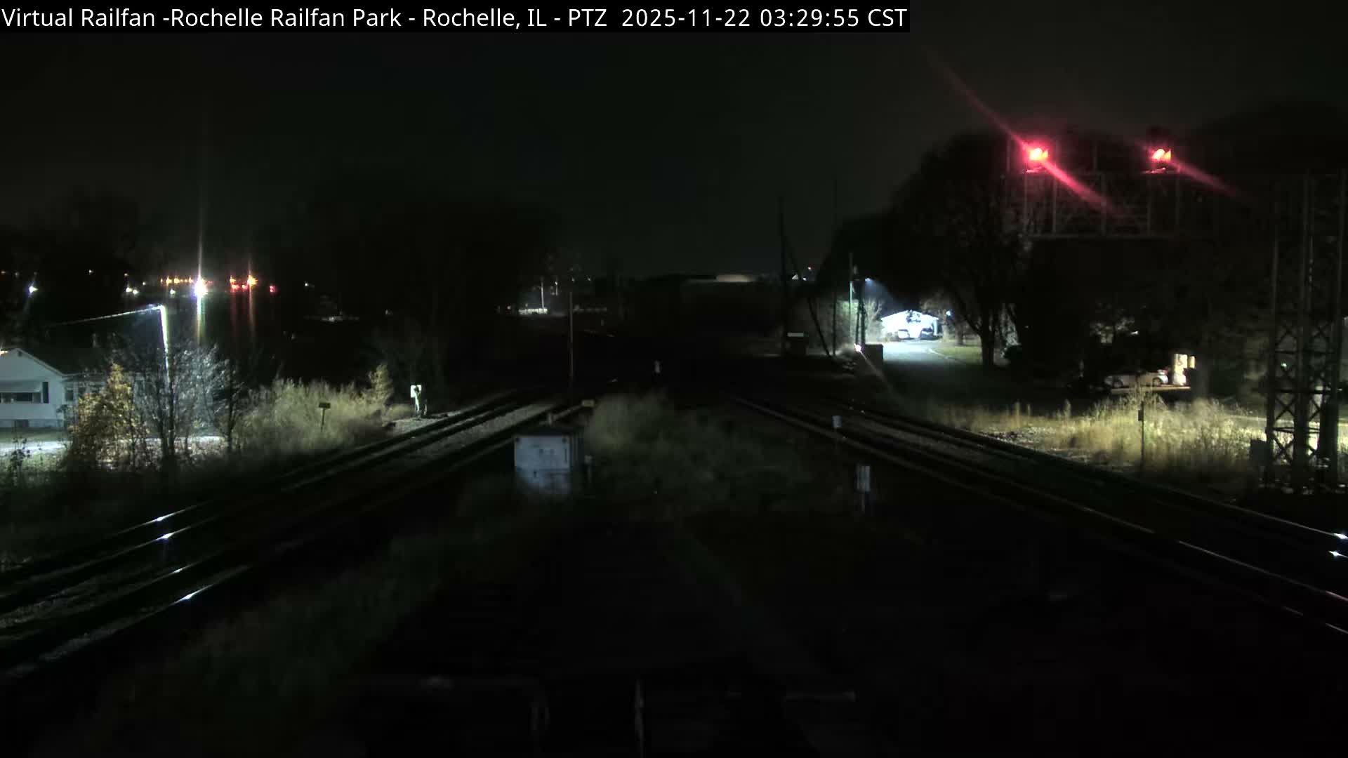 Multiple railway tracks converge through a dark, clear night scene, illuminated by distant town lights and two bright red signal lights on an overhead gantry.