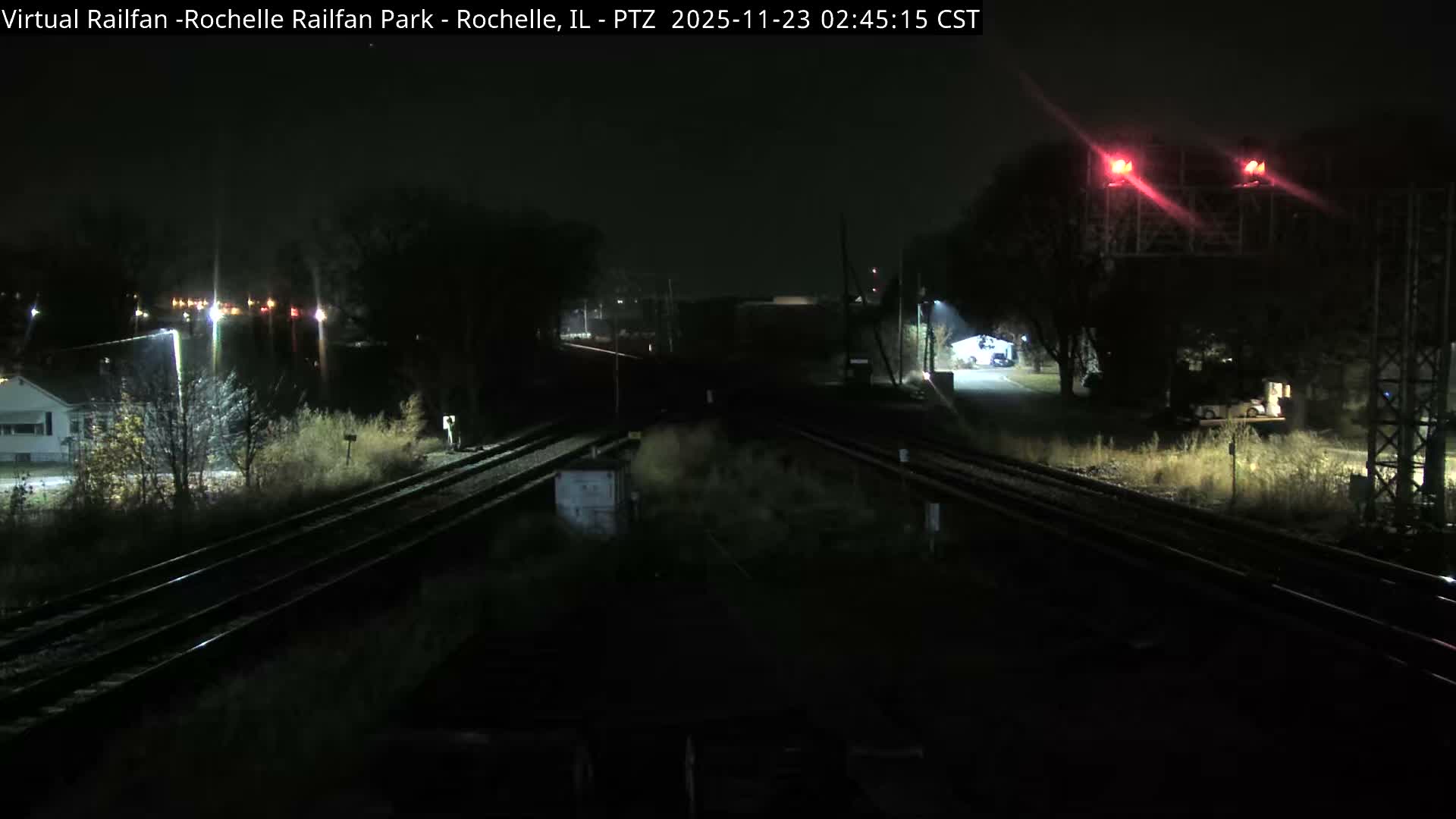 A clear night reveals multiple railroad tracks receding into the distance, bordered by illuminated trees, scattered streetlights, a house on the left, and prominent red railway signals on the right.