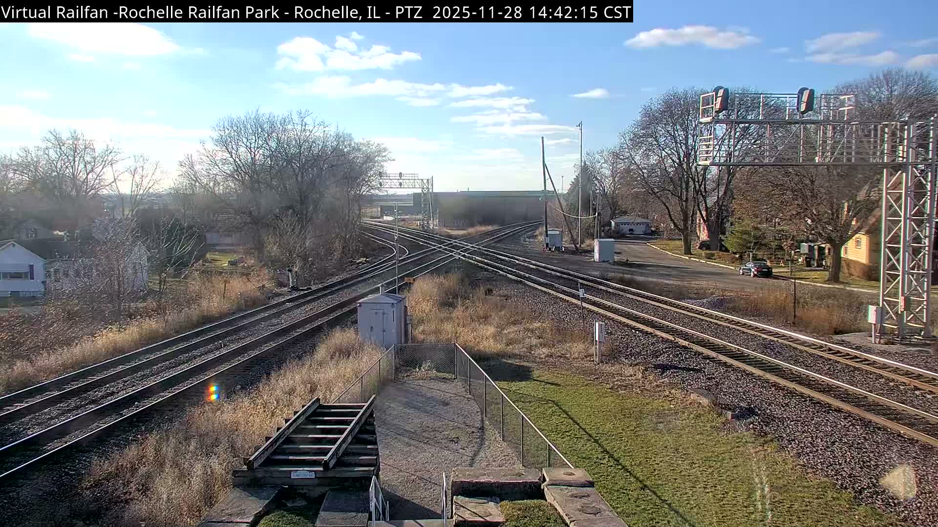 The image displays a sprawling railway junction with numerous tracks intersecting under a large signal bridge, bordered by bare trees, grassy embankments, and houses, all under a bright, sunny sky with scattered white clouds.