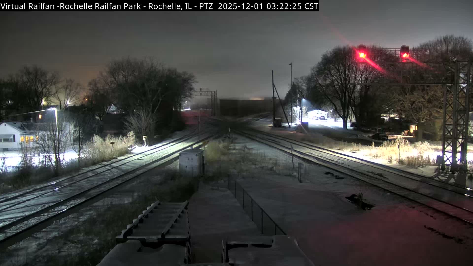 A wintry night scene at a railroad intersection shows snow-dusted tracks, bare trees, and distant houses illuminated by streetlights and active red signal lights under a cloudy sky.