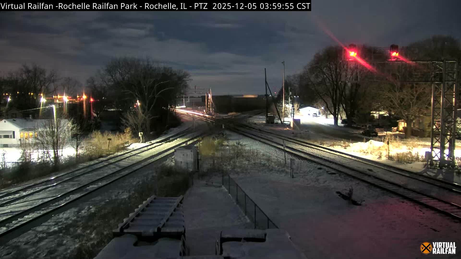 The image depicts a snow-covered railway junction at night, illuminated by numerous artificial lights, including bright red signal lights, with bare trees and distant buildings under a dark, cloudy sky.