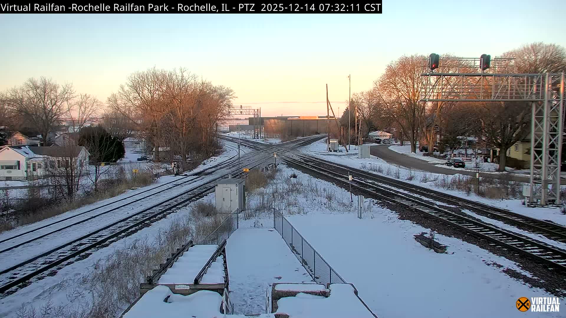 The image depicts a snow-covered railway junction at night, illuminated by numerous artificial lights, including bright red signal lights, with bare trees and distant buildings under a dark, cloudy sky.