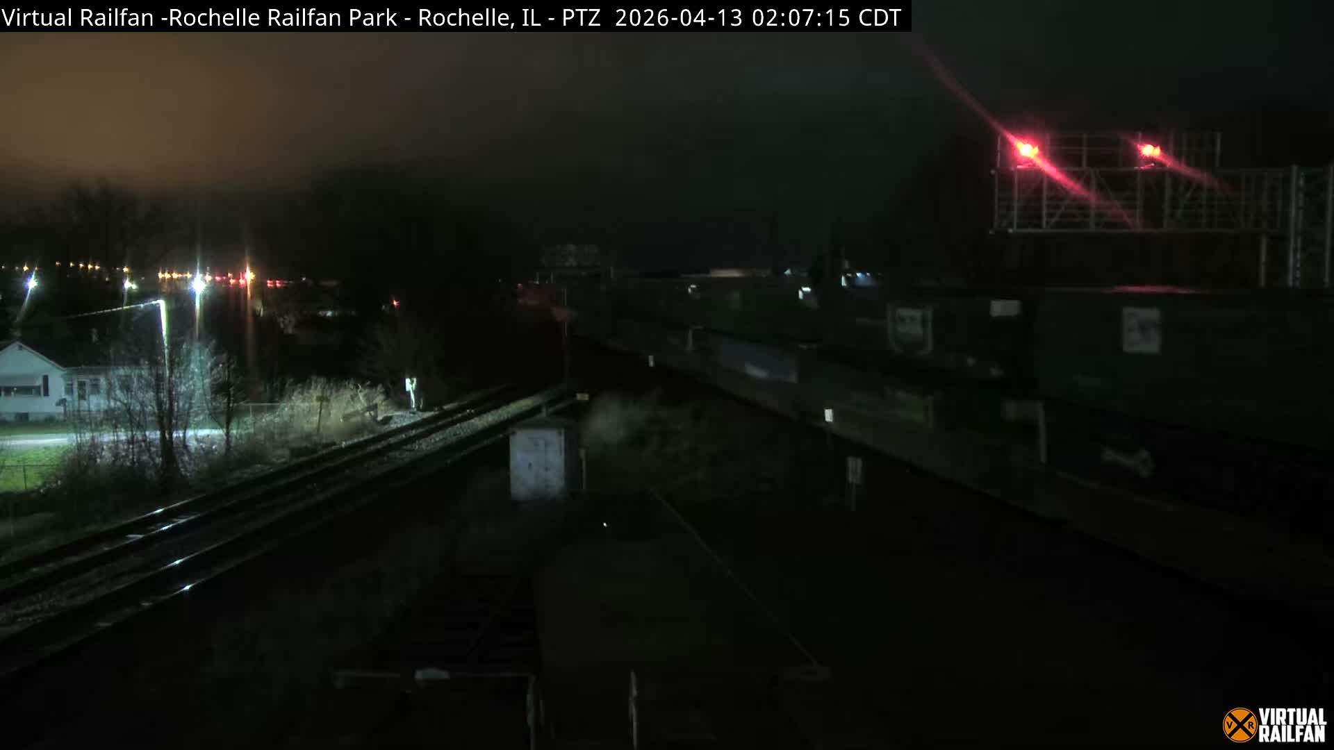 The image depicts a snow-covered railway junction at night, illuminated by numerous artificial lights, including bright red signal lights, with bare trees and distant buildings under a dark, cloudy sky.