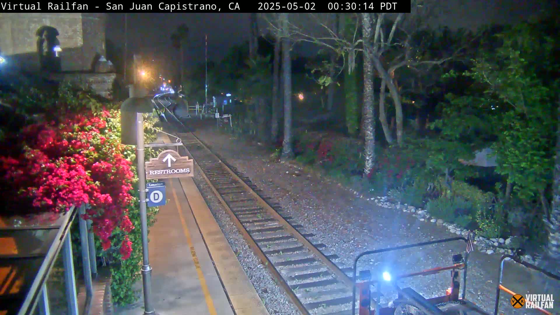 A nighttime view of a train platform with a restroom sign, vibrant red flowers, and railroad tracks extending into a dark, vegetated area.