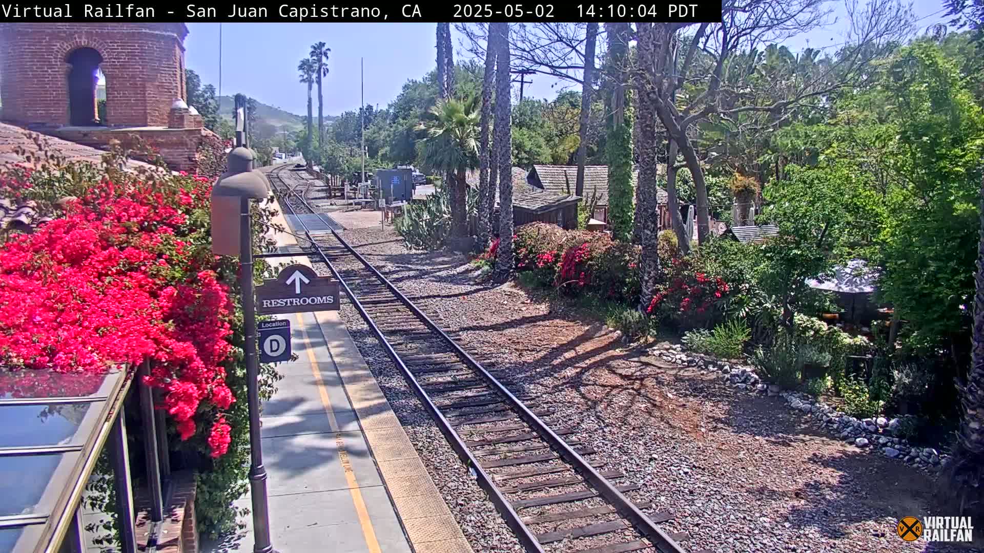 A sunny train station platform with vibrant red flowers, railroad tracks, and lush green landscaping.