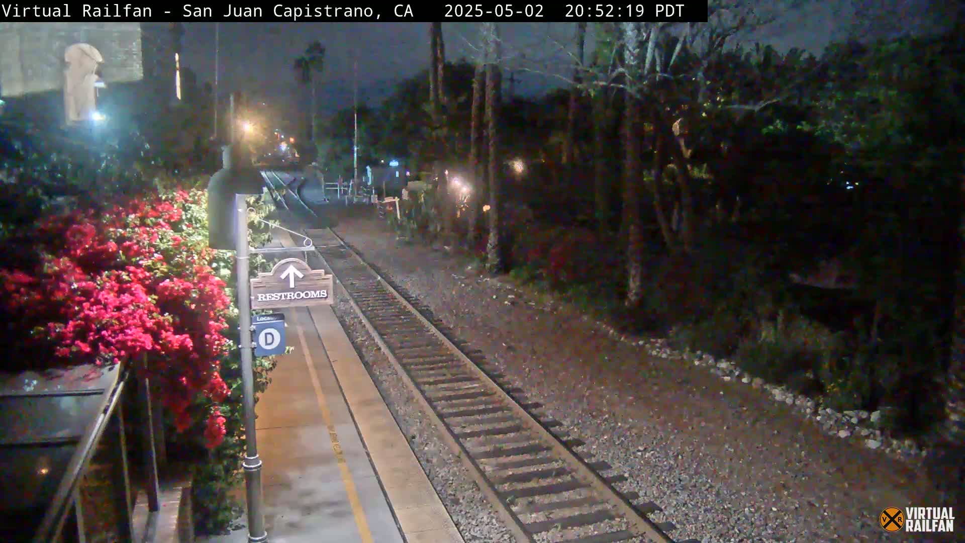 A nighttime view of a train platform with a restroom sign, vibrant red flowers, and train tracks extending into a dark, tree-lined area.