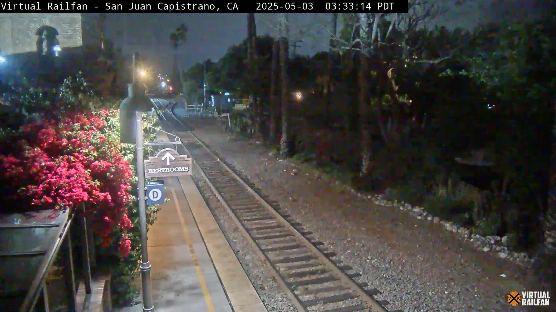 A nighttime view of a train station platform with a restroom sign, blooming pink flowers, and train tracks extending into a dark, tree-lined area.