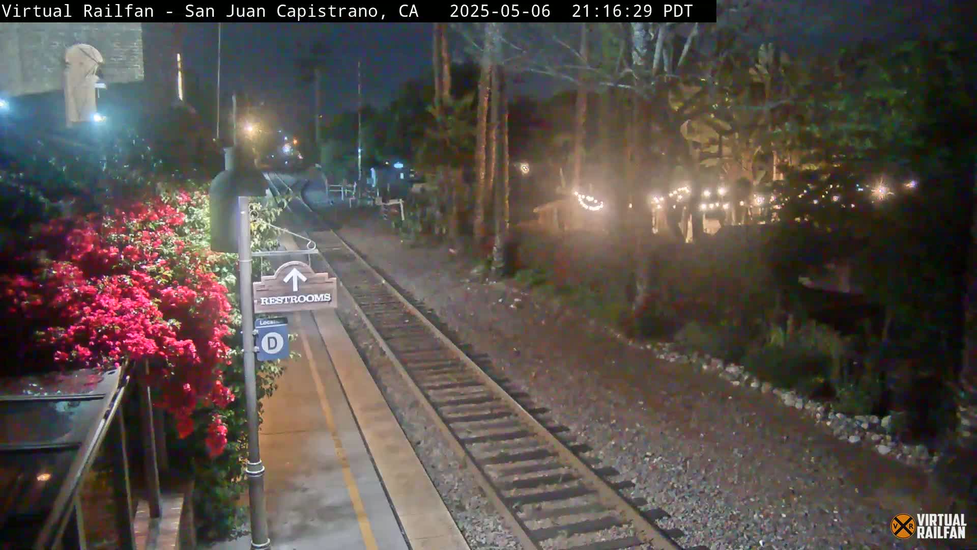 A nighttime view of a train platform with red flowers, a restroom sign, and train tracks leading to a dimly lit area with string lights.