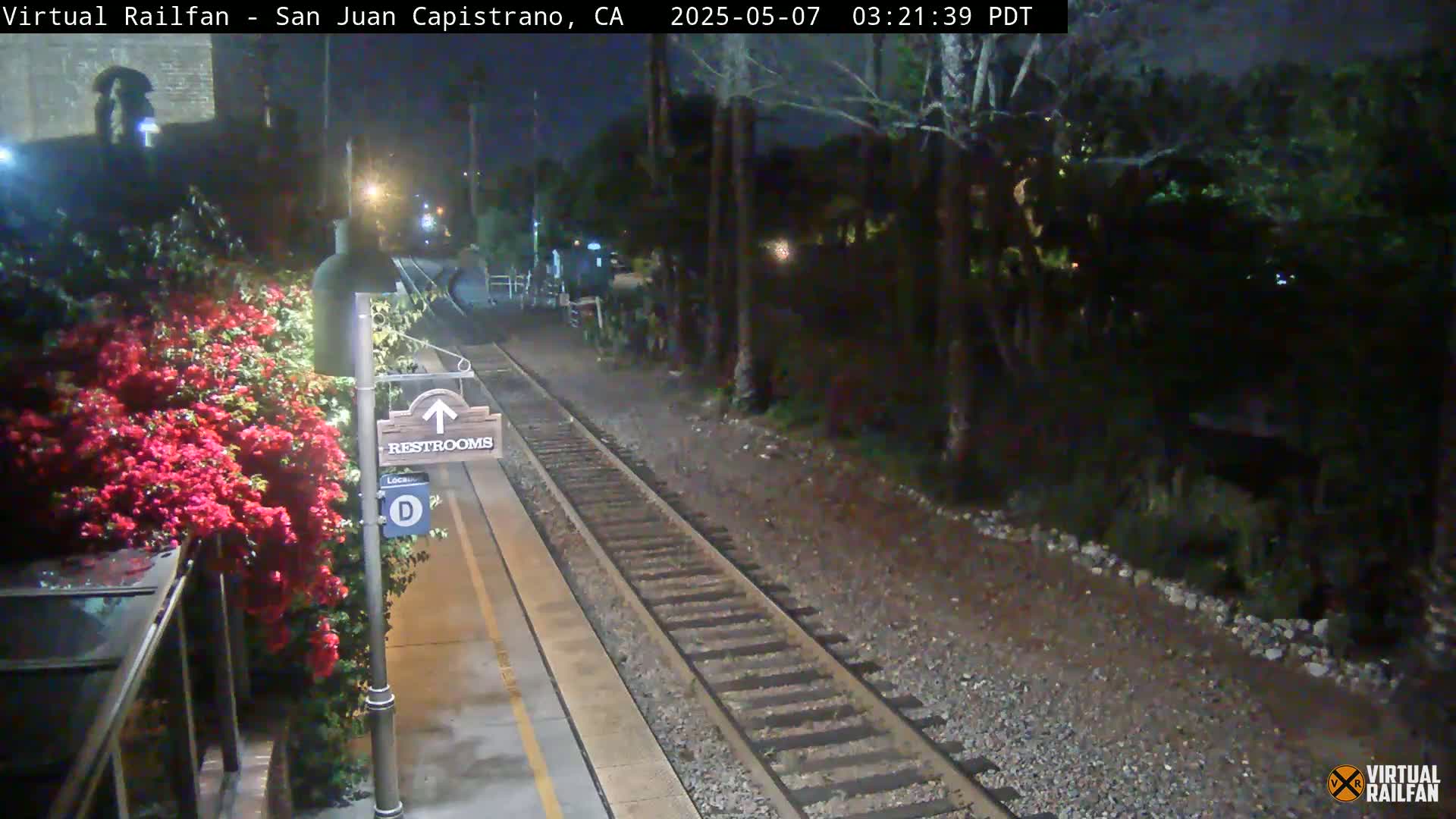 A nighttime view of a train platform with a restroom sign, vibrant pink flowers, and train tracks extending into a dark, tree-lined area.