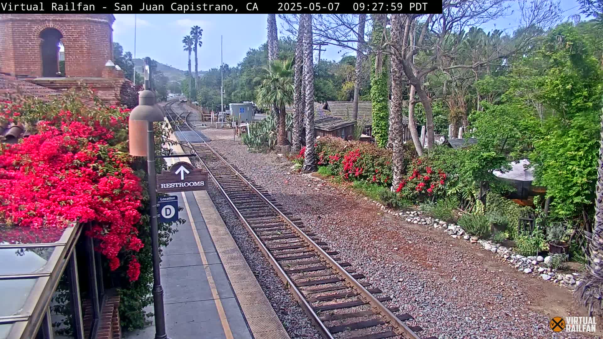 A sunny train station platform with vibrant red bougainvillea, railroad tracks, and lush landscaping.