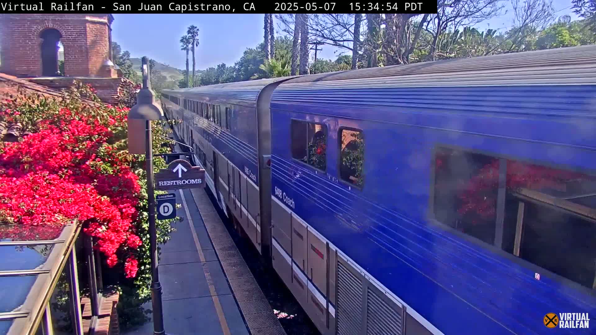 A long blue passenger train sits at a station next to vibrant pink bougainvillea on a sunny day.