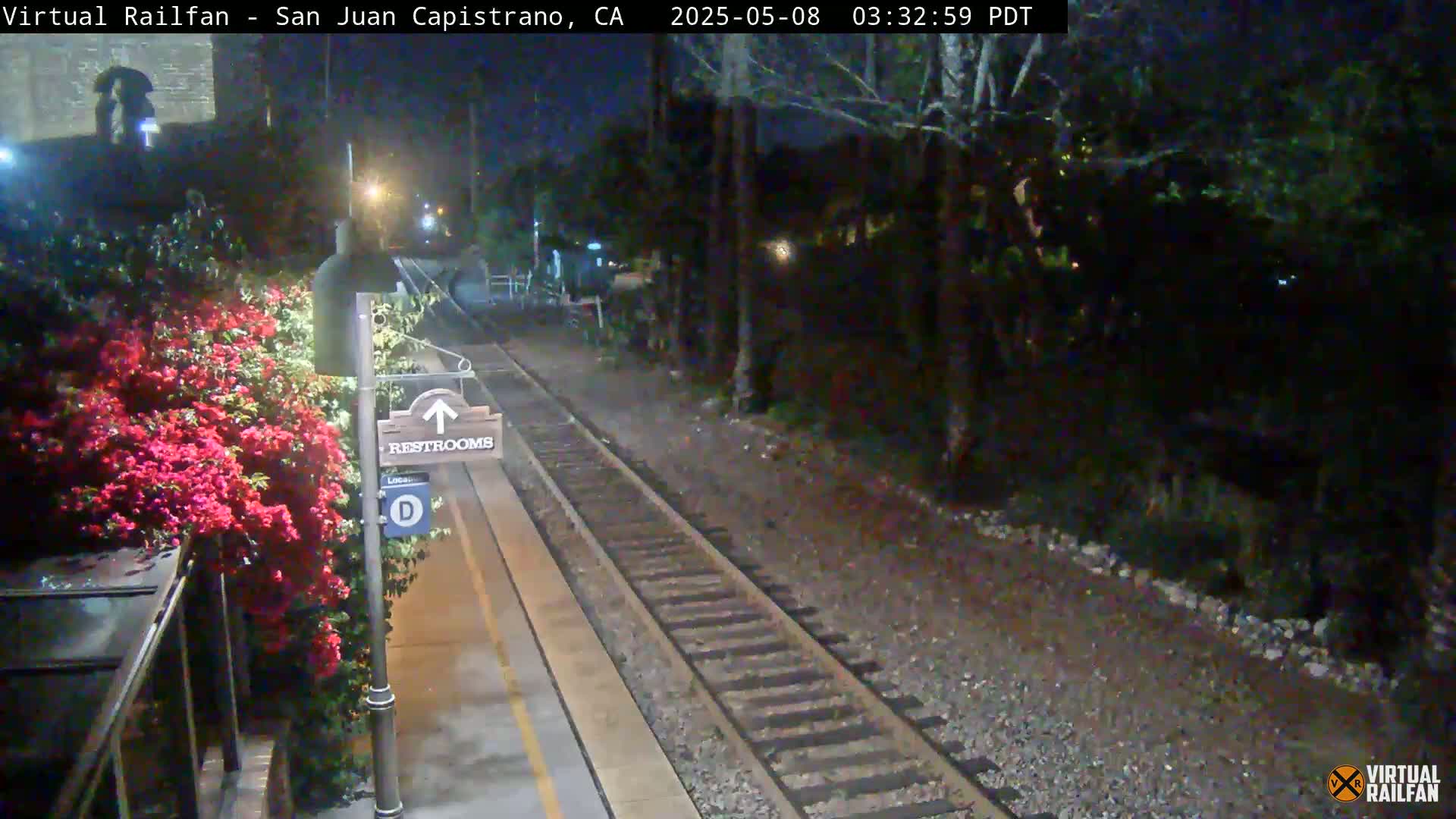 A nighttime view of a train platform with a restroom sign and vibrant pink flowers beside it, showing train tracks extending into a dark, tree-lined area.