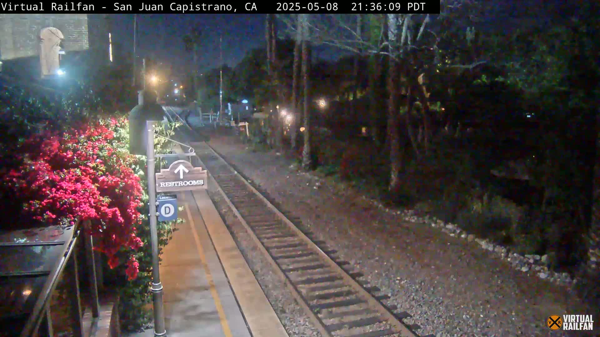 A nighttime view of a train station platform with a restroom sign, lush vegetation, and train tracks extending into the darkness.