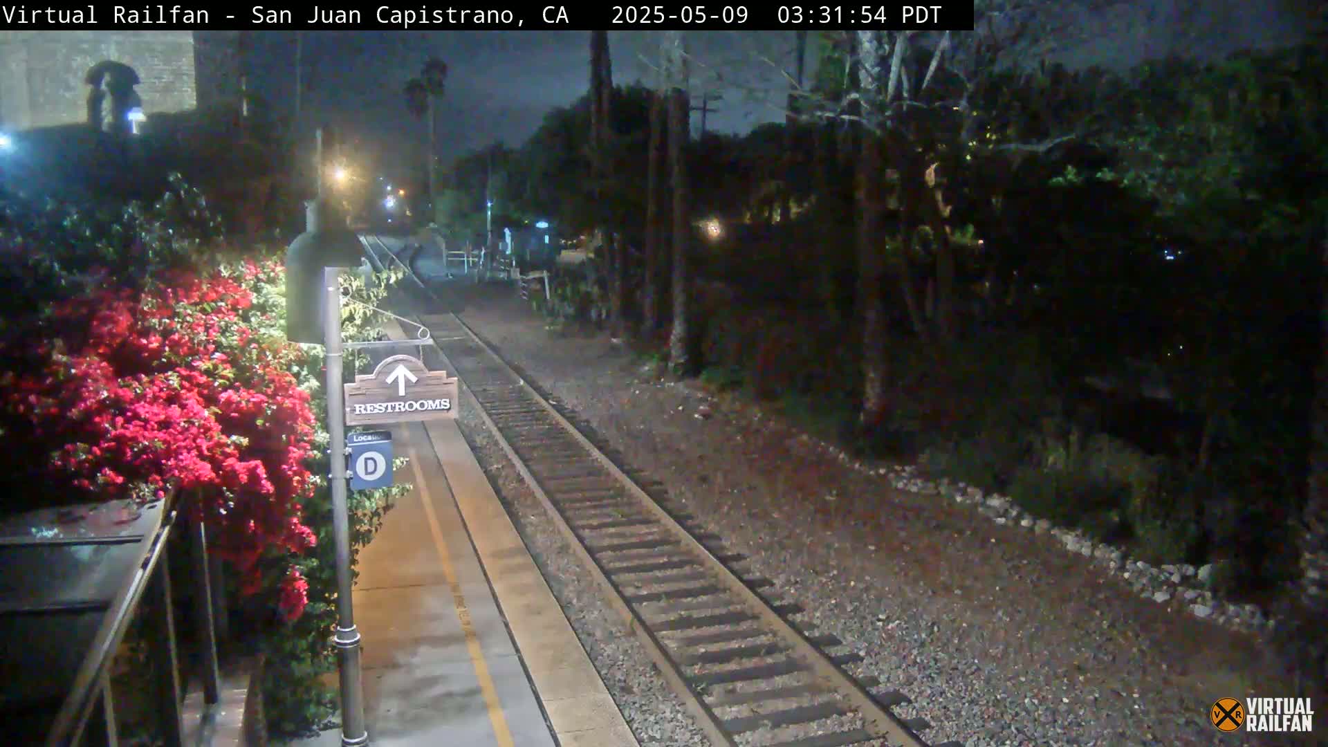 A nighttime view of a train platform with a restroom sign, blooming red flowers, and train tracks extending into a dark, wooded area.