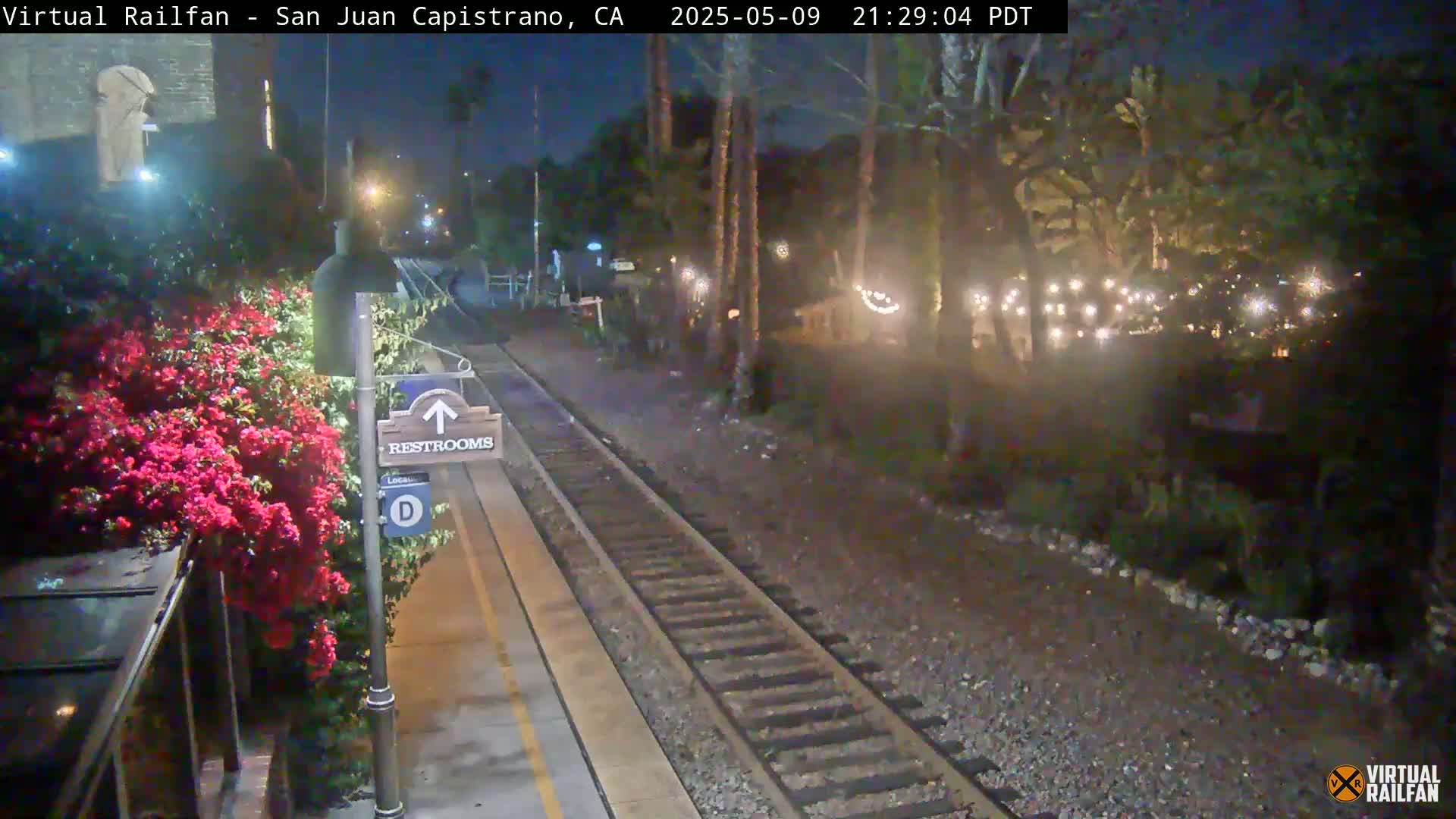 A nighttime view of a train platform with a restroom sign, vibrant red flowers, and string lights illuminating trees in the background.