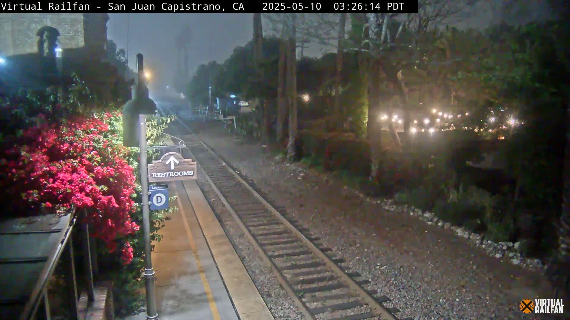 A nighttime train platform with a restroom sign, vibrant pink flowers, and a foggy atmosphere.
