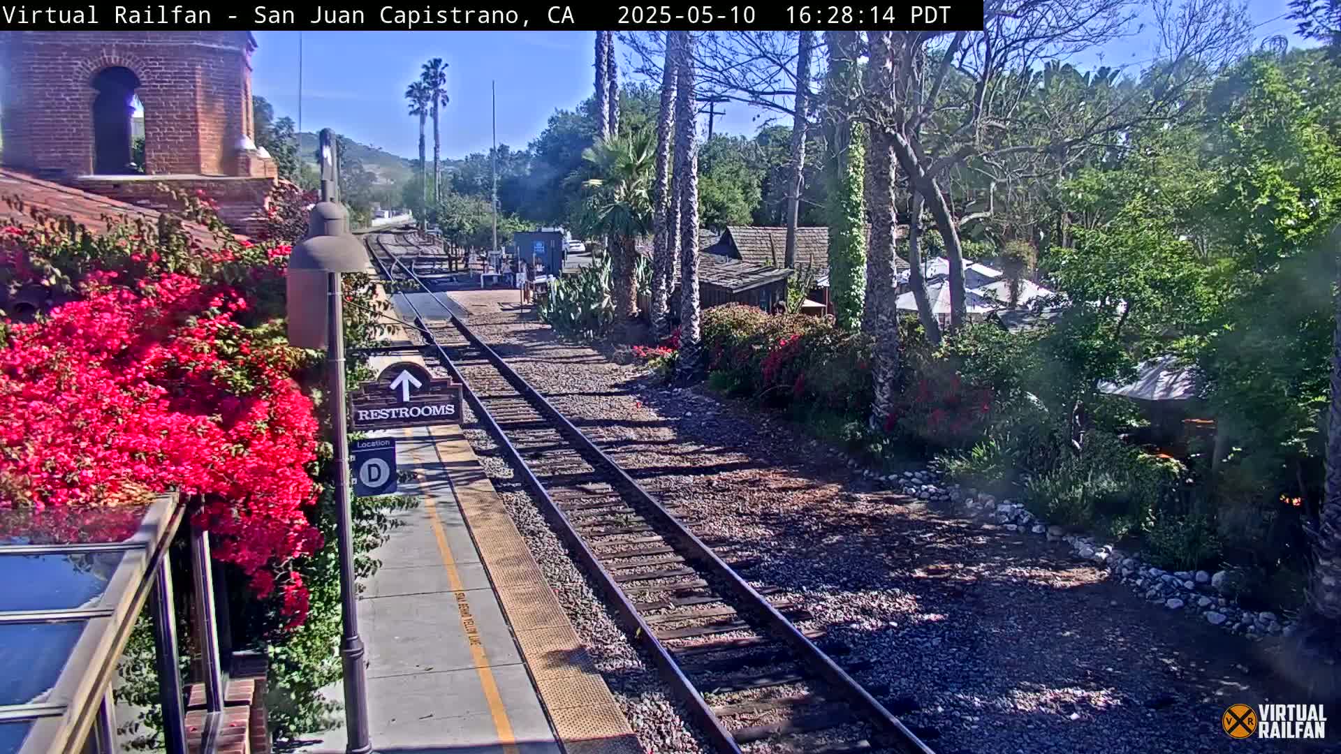 A sunny outdoor scene shows a train station platform with vibrant pink flowers, railroad tracks, and surrounding vegetation.