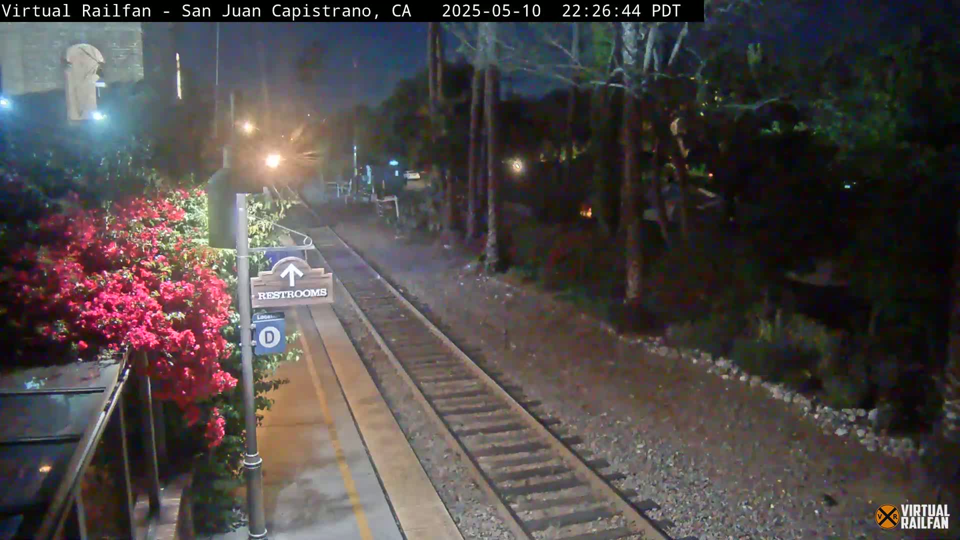 A nighttime view of a train station platform with railroad tracks, a restroom sign, and blooming red flowers next to a covered waiting area.